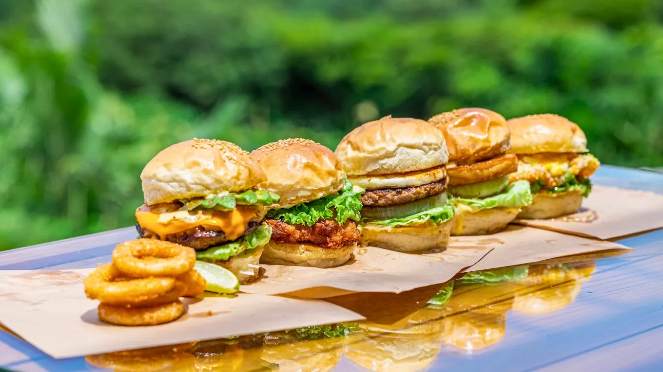 Five burgers and onion rings on a wooden table with greenery behind