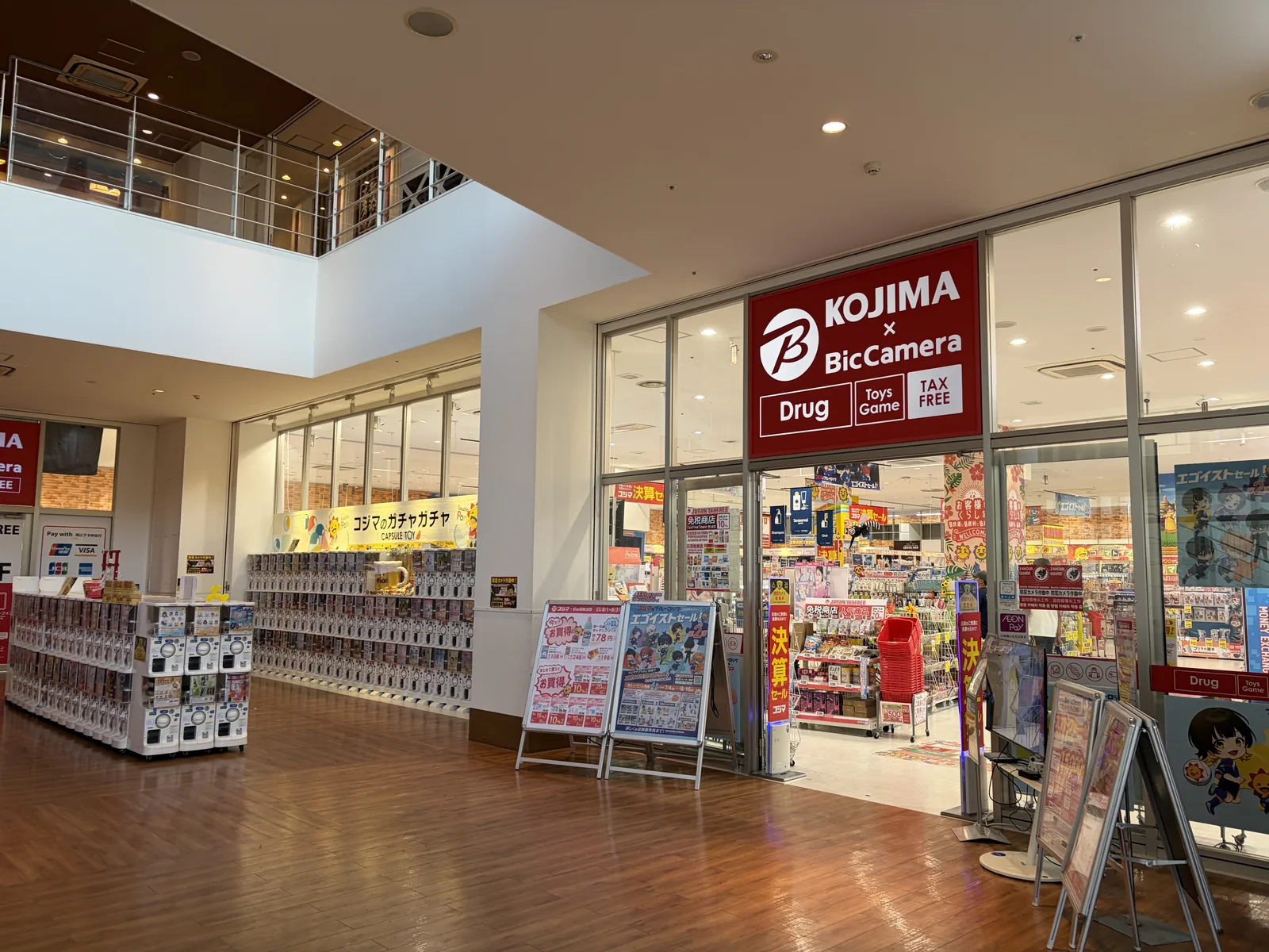 購物中心內 Kojima 與 BicCamera 的店面，招牌上寫著 Drug、Toys Game 與 Tax Free